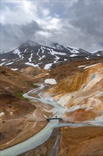 Tourist on bridge over steaming stream between colourful rhyolite mountains with snowfields,