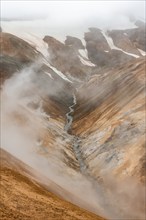 Steaming stream between colourful rhyolite mountains and snowfields, Hveradalir geothermal area,