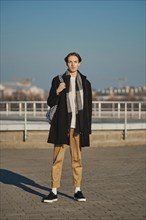 Full length portrait of young man in old wool coat, scarf and short trousers with backpack outdoor