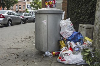 Overfull rubbish bin with trash around and beer cans piled on top in city street