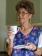 Granny with old smock apron, glasses and wig in the kitchen, holding coffee cup, Germany, Europe