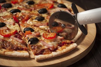 Pizza cutter wheel slicing ham pizza with (capsicum) and olives on wooden board on table