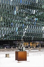 Bronze sculpture of the man with the cello, behind it the glass facade of the Harpa concert hall