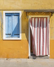 Yellow house facade with entrance door with red and white curtain and window with blue shutters,