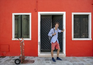 Young man with rucksack standing at a front door, red house facade, colourful houses on the island