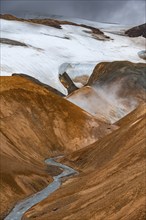 Steaming stream between colourful rhyolite mountains with snowfields, Hveradalir geothermal area,