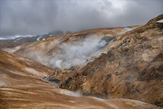 Steaming stream between colourful rhyolite mountains and snowfields, Hveradalir geothermal area,