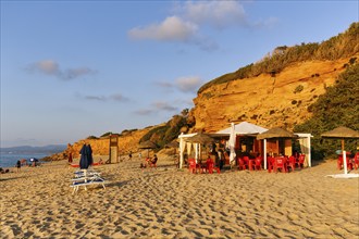 Beach café, red rocks illuminated by the evening sun, sandy beach with tourists, holiday by the