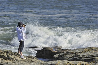 Tourist taking pictures of waves crashing on the rocks at the Pointe Saint-Gildas, Saint Gildas