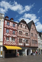 Medieval half-timbered houses in red and white with shops and passers-by under a clear sky,