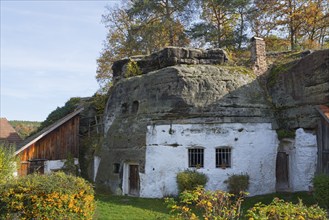 A rustic house integrated into a rock face, surrounded by autumn vegetation, rock dwelling, museum,