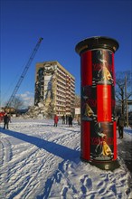 People walk past an advertising pillar while a building is being demolished in the background