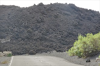 A road ends in front of a large accumulation of black volcanic rock and some vegetation, last