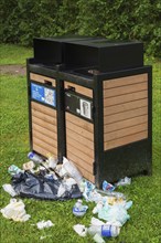 Black painted metal and natural wood garbage recycling bins with discarded thrash on green grass