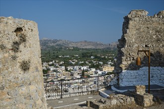 View through a ruined wall to a village in the distance, surrounded by mountains and clear skies,