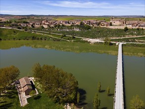 An aerial view of a river with a bridge leading to a village with many houses, surrounded by