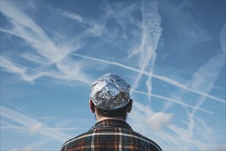 Chemtrail conspiracy theory concept with man with tinfoil hat looking at sky with condensation