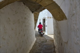 Motorcyclist riding through a narrow alley with whitewashed walls on a sunny day, Chora, main town