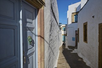 A narrow alley with white buildings, blue doors and wooden windows under a clear blue sky, Chora,