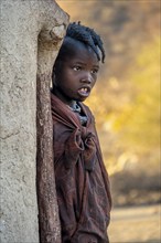 Himba girl leaning against a traditional mud hut, in the morning, traditional Himba village,