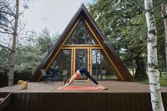 Flexible woman standing in dog's face-down pose on wooden terrace of her summer cabin