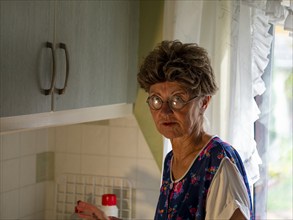 Granny with old smock apron, glasses and wig at the kitchen window in the kitchen, Germany, Europe