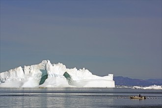 Small fishing boat surrounded by seabirds in front of iceberg with large ice cave, Ilulissat, Disko