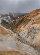 Steaming stream between colourful rhyolite mountains and snowfields, Hveradalir geothermal area,