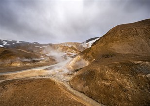 Steaming stream between colourful rhyolite mountains with snowfields, Hveradalir geothermal area,