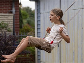 Boy swinging while sitting on children's swing at shed and garden gate, Germany, Europe