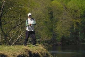 Fisherman throwing the bait into river standing on river bank in sunny day with green forest in