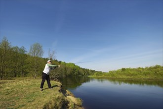 Fisherman with a spinning rod catching fish on a river at sunny summer day with green trees at