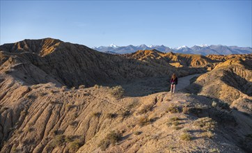 Tourist standing in a landscape of eroded hills at sunrise, Badlands, white mountain peaks of the