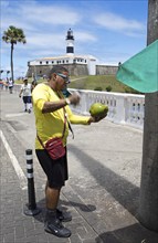 Brazilian man opening a coconut, Faro da Barra lighthouse in the back, Salvador, State of Bahia,