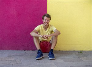 Young man sitting in front of a house wall, with trousers and T-shirt in the same colours as the