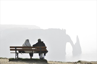 Elderly tourists sitting on bench looking at the Porte D'Aval, a natural arch in the chalk cliffs