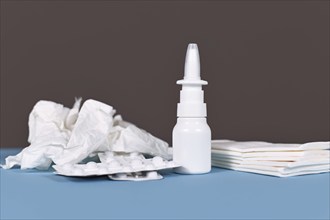 White bottle with nasal spray with crumbled tissues and pills used against common cold on blue