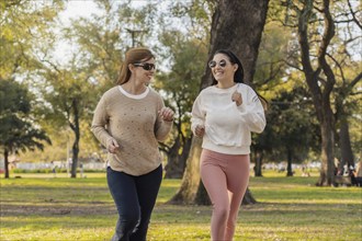 Two women run side by side in a lush green park on a sunny day. They enjoy the outdoors, staying