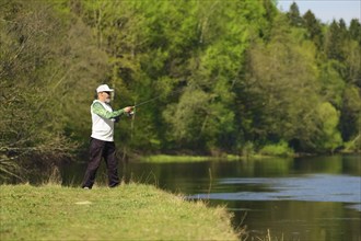 Fisherman with a spinning rod catching fish on a river at sunny summer day with green trees at