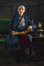 Attractive lady in oldfashioned clothes reading ancient book near vintage kerosene lamp