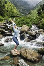 Young sporty fit woman doing yoga asana Utkatasana (chair pose) outdoors at tropical waterfall