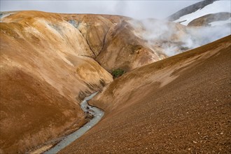 Steaming stream between colourful rhyolite mountains and snowfields, Hveradalir geothermal area,