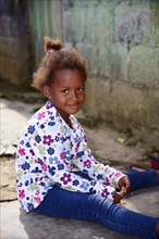 A smiling child with a floral print top sits casually in front of a house, between Pedro Sanchez