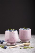 Yoghurt with blueberry and sesame in a glass and wooden spoon on gray and black background. side