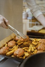 Closeup view of lifting meat patty from baking tray with potato wedges with kitchen tongs