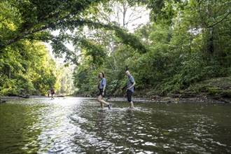 Young woman and man in the tropical rainforest crossing a stream, tourists wading through a stream,