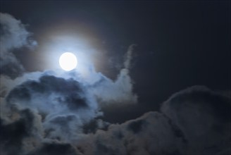 The full moon shines brightly in a cloudy, stormy, black sky and has a halo in rainbow colours