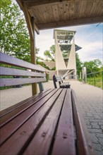 A wooden bench next to a hiking trail offers a view of a large modern observation tower,