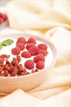 Yogurt with raspberry and goji berries in ceramic bowl on white concrete background and orange