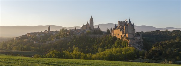 Panoramic view of a historic castle and town surrounded by green fields and mountains at sunrise,